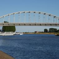 Witte boogbrug John Frostbrug over brede rivier in Arnhem met passagiersschepen eronder en groene bomen langs de oevers onder heldere hemel.