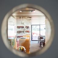 Zicht door ronde patrijspoort op helder interieur van MS Ave Maria met houten balie, planken, eethoek en glazen toegangsdeur.