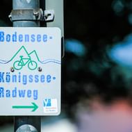 Wit richtingbord voor Bodensee-Königssee fietsroute met fietspictogram, berggrafiek en pijl, gemonteerd op een paal.