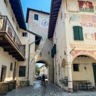 Historisch plein in Friuli met klokkentorenboog en gebouw versierd met kleurrijke fresco's. Groene luiken en buitenstoelen zichtbaar.