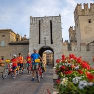 Groep fietsers met fietsen voor de middeleeuwse stenen poort van kasteel Sirmione. Rode bloemen op de voorgrond.
