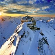 Schilthorn bergstation op besneeuwde top bij zonsopgang, omringd door besneeuwde Alpentoppen onder dramatische bewolkte hemel.