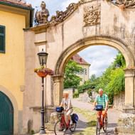 Twee fietsers rijden door een sierlijke barokke poort met beelden bij Schloss Horneck in Gundelsheim. De verweerde stenen poort heeft decoratieve sculpturen.