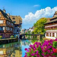 Traditionele vakwerkhuizen langs een kanaal in Petite France, Straatsburg, met kleurrijke bloemen op de voorgrond en blauwe lucht.