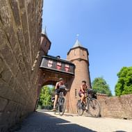 Twee fietsers rijden door een bakstenen poort met torens bij Kasteel de Haar nabij Utrecht op een zonnige dag met blauwe lucht.