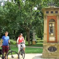 Twee fietsers met fietsen bij een sierlijke historische klokkentoren in het kuurpark van Franzensbad, omringd door groene bomen.
