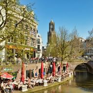 Buitenterrassen langs de Utrechtse gracht met rode parasols. Historische gebouwen langs de waterweg met Domtoren op de achtergrond.