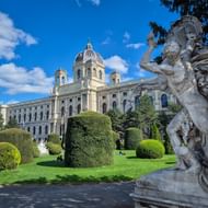 Kunsthistorisches Museum in Wenen met sierlijke koepel en gevel. Verzorgde tuinen met gesnoeide hagen en klassiek standbeeld op voorgrond.