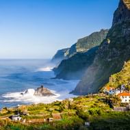 Witte huizen genesteld op groene terrasvormige hellingen onder steile kliffen op Madeira. Golven breken tegen rotsen langs de blauwe Atlantische kustlijn.