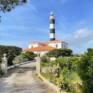 Witte vuurtoren met zwart-witte strepen in Portocolom, Mallorca, omgeven door mediterrane vegetatie en bereikbaar via een verharde weg.