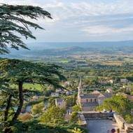 Verhoogd uitzicht op een Provençaals dorp met kerk en stenen gebouwen, omlijst door ceders. Glooiende landbouwvallei strekt zich uit naar bergen.
