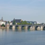 Meerbogige stenen brug over een kalme rivier in Maastricht met historische gebouwen en kerktorens langs de waterkant.