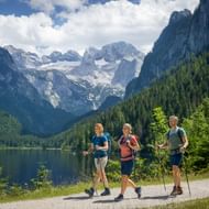 Drie wandelaars met trekkingstokken langs het Gosausee-meer in Salzkammergut. Dachstein-gletsjer en beboste hellingen op de achtergrond.