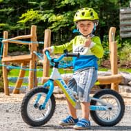 Jong kind met gele helm en groen shirt zittend op blauwe loopfiets op speelplaats met houten klimtoestel op de achtergrond.