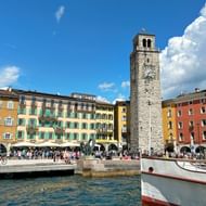 Havenplein in Riva del Garda met middeleeuwse klokkentoren, kleurrijke gebouwen met arcades, aangemeerde boten en toeristen.