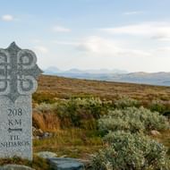 Grijze stenen mijlpaal met '208 KM TIL NIDAROS' en Keltisch kruis in Noorse toendra met bergen op de achtergrond.