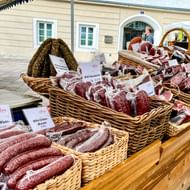 Marktkraam in Zell am See met regionale wildspecialiteiten in rieten manden, waaronder verschillende worsten en vleesproducten met Duitse labels.