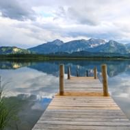 Houten steiger uitstrekkend in kalme Hopfensee met perfecte bergreflecties. Alpentoppen zichtbaar op achtergrond onder bewolkte lucht.