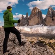Vrouwelijke wandelaar in groene jas met trekkingstokken op rotsachtig terrein, kijkend naar de iconische Tre Cime pieken in de Dolomieten.