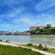 Uitzicht over de Donau naar het Kasteel Museum Linz, een groot barok gebouw op een heuvel, met de stadshorizon en bergen op de achtergrond.