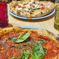 Twee Italiaanse pizza's op blauwe borden met verse basilicum, geserveerd met rode en gele getextureerde glazen op een houten tafel.