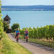 Twee fietsers rijden door wijngaarden bij Meersburg met het Bodenmeer en een historische toren op de achtergrond.
