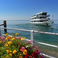 Witte passagiersboot nadert een pier op het Bodenmeer met kleurrijke bloemen op de voorgrond en heldere blauwe lucht erboven.
