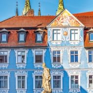 Blauw barok gebouw met wit ornamenteel stucwerk in Bamberg. Een gouden standbeeld staat ervoor. Erachter rijzen kerktorens boven rode daken uit.