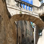 Smalle geplaveide straat in Uzès met een stenen boogbrug die gebouwen verbindt. Beige stenen gevels langs de straat onder een blauwe lucht.