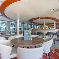 Moderne lounge bar op MS Lisabelle met witte gebogen stoelen, ronde houten tafels, bar met krukken en grote ramen met gordijnen.