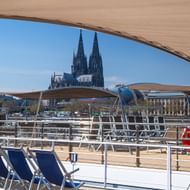 Zonnedek van MS Lisabelle met blauwe ligstoelen en rode reddingsboei, uitzicht op de tweelingtorens van de Keulse Dom over de Rijn.