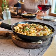 Zwarte pan met Kasnockn-pasta gegarneerd met kruiden en paneermeel op houten plank. Rode wijnglas en tweede pan op de achtergrond.