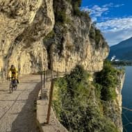 Fietser in geel shirt op Strada del Ponale, een schilderachtig pad uitgehouwen in rotswand boven Gardameer met bergen op de achtergrond.