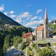 Panoramisch uitzicht op Bad Gastein in een Alpendal met prominente kerktoren, kleurrijke gebouwen en beboste bergen onder blauwe lucht.