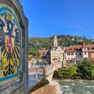 Stenen brug in Merano met mozaïek rijksadelaar embleem. Historische stadsgebouwen en kerktoren zichtbaar over de rivier onder blauwe lucht.