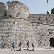 Vier toeristen lopen langs massieve stenen vestingmuren met ronde toren in de Baai van Kotor, Montenegro. De oude verdedigingswerken hebben een boogvormige ingang.