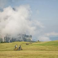 Twee fietsers rijden door groene weiden in de Dolomieten met dramatische bergtoppen gedeeltelijk bedekt door wolken en houten hutten in de verte.