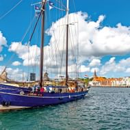 Blauw tweemast zeilschip in haven van Sønderborg met kleurrijke waterkantgebouwen en kerktoren op achtergrond onder blauwe lucht.