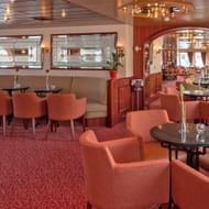 Elegante salon bar op MS Switzerland met koraalkleurige fauteuils, ronde tafels en lange eettafel. Met rood tapijt en warme verlichting.
