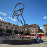 Grote metalen sculptuur van een gestileerde figuur in een rond fonteinbekken op een stadsplein in Alba, Piemonte, met historische gebouwen op de achtergrond.