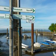 Bewegwijzering voor Elbe fietsroute met afstanden naar Elmshorn, Glückstadt, Neuendorf en andere bestemmingen, met jachthaven en boten op achtergrond.