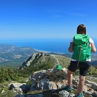 Vrouwelijke wandelaar met groene rugzak staat op rotsachtige bergtop met uitzicht op Ligurische kust met blauwe Middellandse Zee.