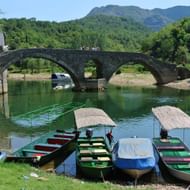 Historische stenen boogbrug over groene rivier in Montenegro met boten op voorgrond en beboste bergen op achtergrond.