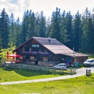 Huberhütte berghut met rode parasols op groene Postalm weide, omgeven door dicht naaldbos en geparkeerde auto's.