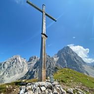 Houten topkruis op de Sulzenschneid met dramatisch Dachstein-gebergte op de achtergrond onder blauwe hemel met witte wolken.