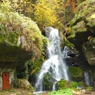 Lichtenhainer waterval stroomt over met mos bedekte rotsen in herfstbos. Rode houten structuur zichtbaar aan linkerkant.