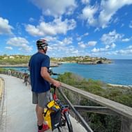 Fietser in blauw shirt en helm staat met fiets op kustpad met uitzicht op turquoise Middellandse Zee en Porto Cristo kustlijn.