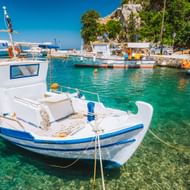 Wit-blauw vissersboot afgemeerd in kristalhelder turquoise water van een Kretenzische haven, met andere traditionele boten en groene heuvel op de achtergrond.
