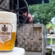 Glazen bierpul Svitavy met schuimkraag op witte tafel in brouwerijgaard. Historische brouwapparatuur zichtbaar op achtergrond tussen groen.