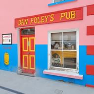 Dan Foley's Pub aan de Dingle Way Ierland Kleurrijke Ierse pub gevel met roze en blauwe muren. Rood bord 'Dan Foley's Pub' boven rode deur. Raam toont Guinness bord.